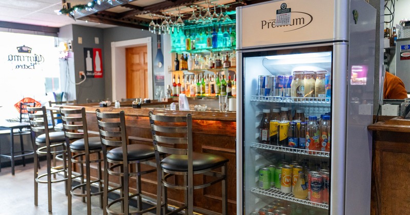 Bar interior with wooden counter, metal chairs, liquor shelves and a white beverage fridge.