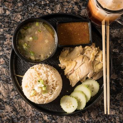 Hainanese chicken rice with soup, chili sauce, and cucumbers on a black plate.