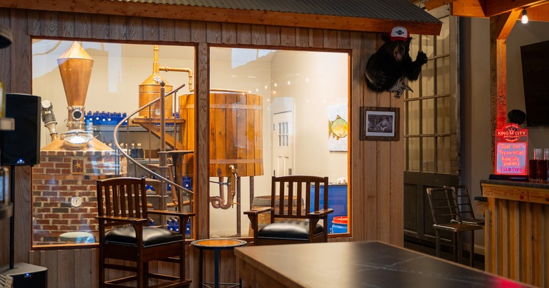 Two upholstered chairs and a table, view of a distillery room behind the glass