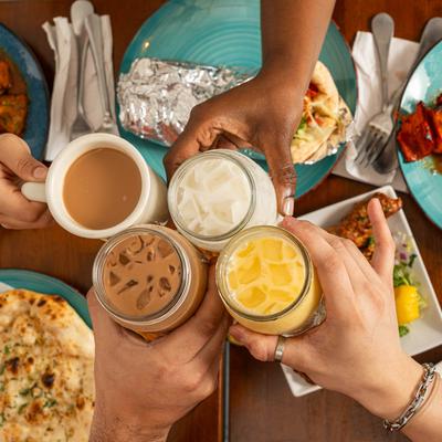 People holding various drinks clink glasses over a table filled with colorful Indian dishes.
