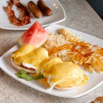 Eggs Benedict topped with hollandaise sauce, served with hash browns and watermelon slice.