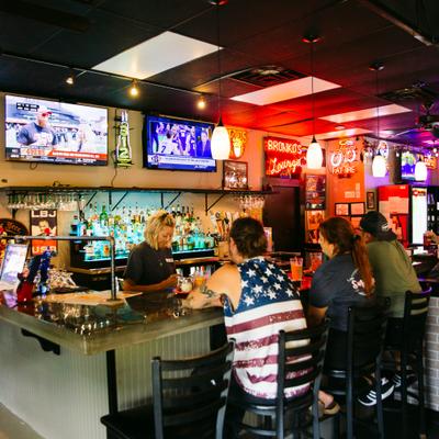 Interior of a bar featuring a blonde bartender, patrons at the bar and colorful bottles.