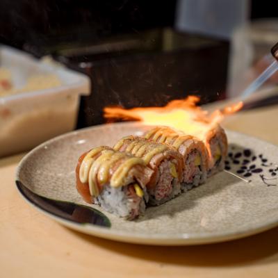 A chef uses a blowtorch to sear a saucy sushi roll.