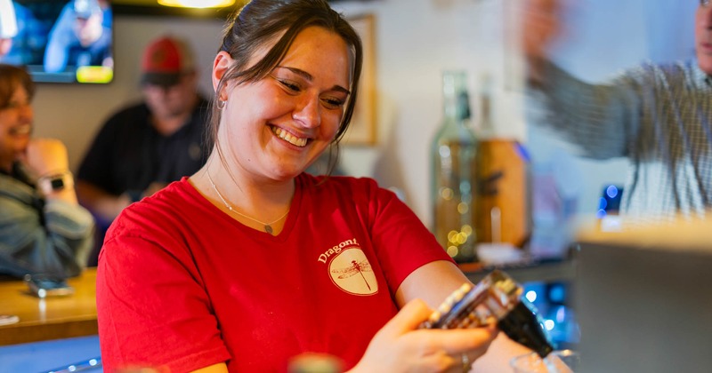 Smiling employee in a Dragonfly red shirt pours drinks at a lively bar