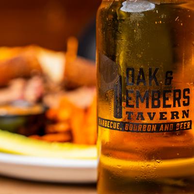 Selective focus on a beer glass with Oak and Embers trademark, a dish in background.