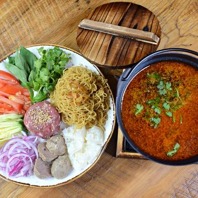 A plate of noodles, meats, and vegetables next to a bowl of broth on a wooden table.