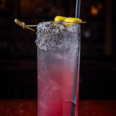 Fizzy purple cocktail in a highball glass garnished with dried lavender buds and lemon peel.