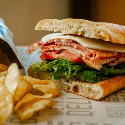 Close up of a sandwich with cured meat, cheese, tomato, and lettuce, served with chips.