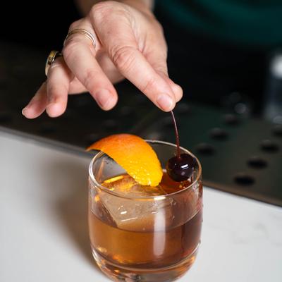 A bartender garnishes Barrel Aged Black Manhattan with lemon peel and a dark cherry.