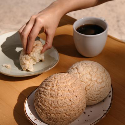 A hand breaking concha pastry beside a cup of black coffee.