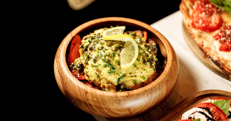 A wooden bowl holds creamy chicken francese topped with lemon and capers