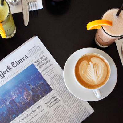 A cup of latte with leaf art beside a newspaper and fresh-squeezed juices.