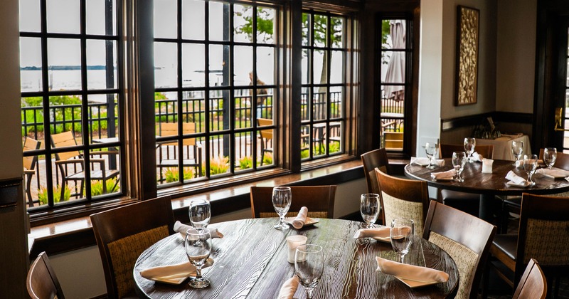 A dining room with large windows overlooking a patio and a lake, featuring round wooden tables