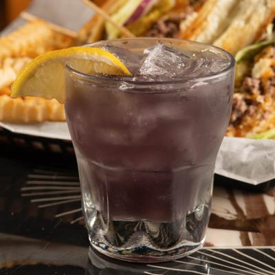 A purple cocktail on the rocks with a lemon slice, food in the background.