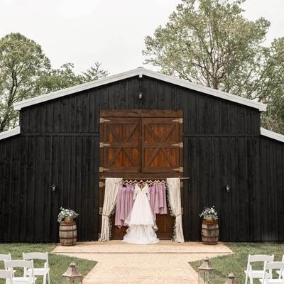 Wedding dress hung in front of a rustic barn with chairs and floral decorations nearby.