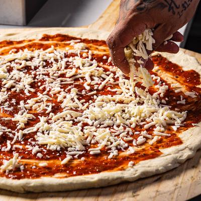 A hand sprinkling shredded mozzarella cheese over pizza dough.
