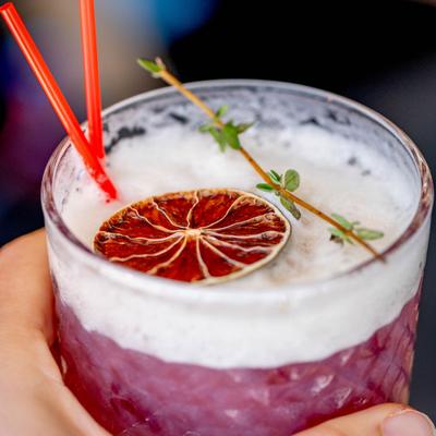 Hand holding a Thonglor Sour cocktail, garnsihed with dried lime wheel and thyme.