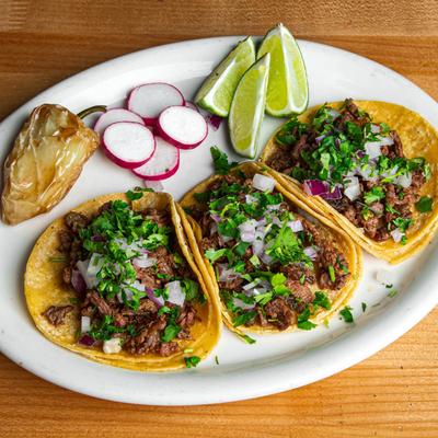 Street tacos with onions and cilantro, served with lime wedges, radishes, and roasted pepper.