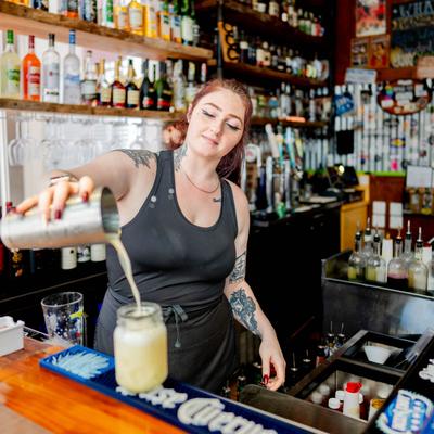 A bartender pouring a cocktail drink into a mason jar on a bar counter.
