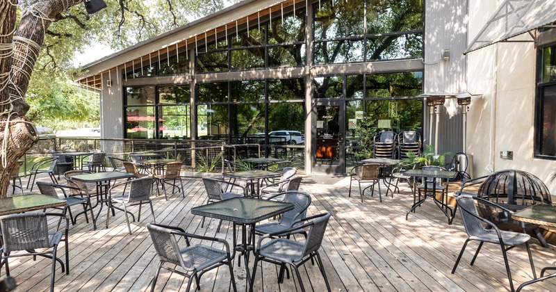An outdoor restaurant deck features numerous tables and chairs under a large tree