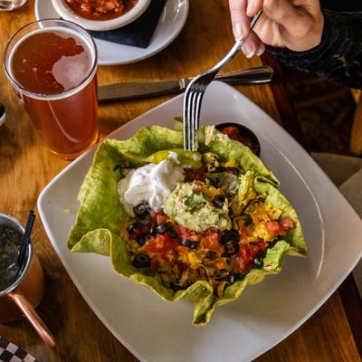 A hand holding a fork over a taco salad beside a glass of beer.