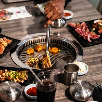 Hand using metal tongs to flip meat on an interactive Korean BBQ grill.