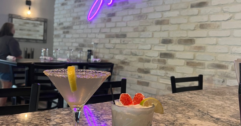 Two cocktails sit on a granite bar in front of a purple neon sign reading "Sophia's"