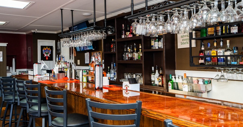 A bar with a wooden counter, bar stools, hanging wine glasses and shelves of liquor bottles