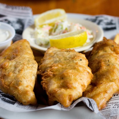 3 pieces of fried fish on top of chip style french fries on a plate with a side order of coleslaw, lemon and tartar sauce