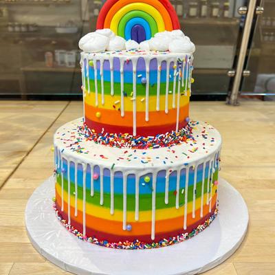 Two-tiered cake with rainbow stripes, white icing drips, colorful sprinkles, and a rainbow topper.