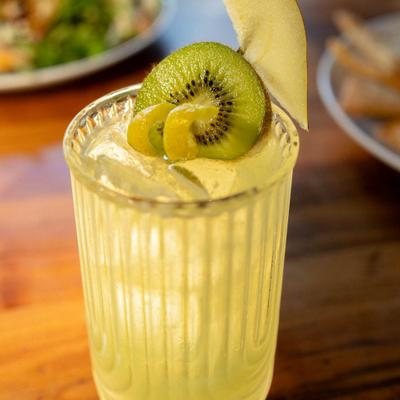 Cocktail in a long glass, garnished with lemon leel and slices of pear and kiwi.