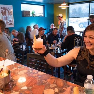 A guest raises a cocktail at the bar counter, diners are seated at tables in the background.