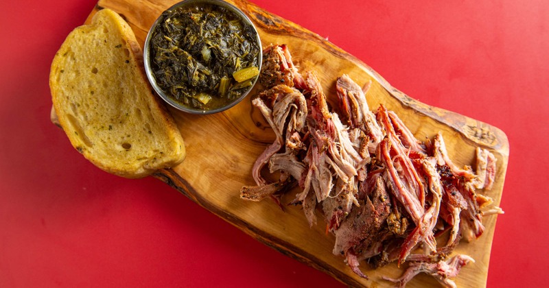 A wooden board with pulled pork, collard greens, and Texas toast