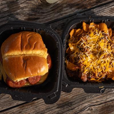 BBQ sausage sandwich and cheesy Frito pie in black takeout containers on a wooden table.