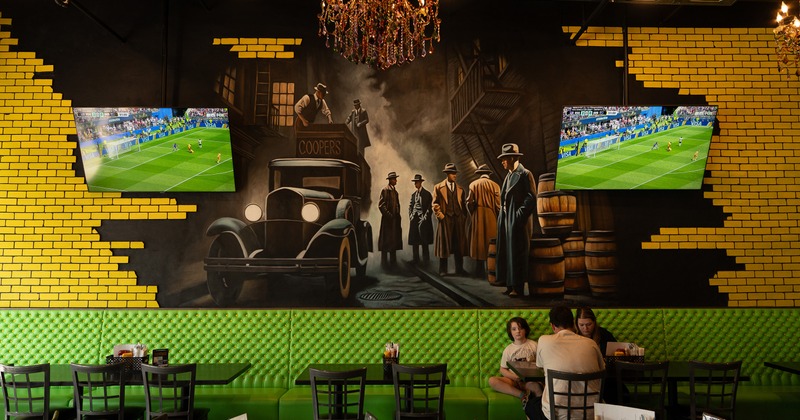 A vintage-style mural of mobsters and cars sits behind patrons inside