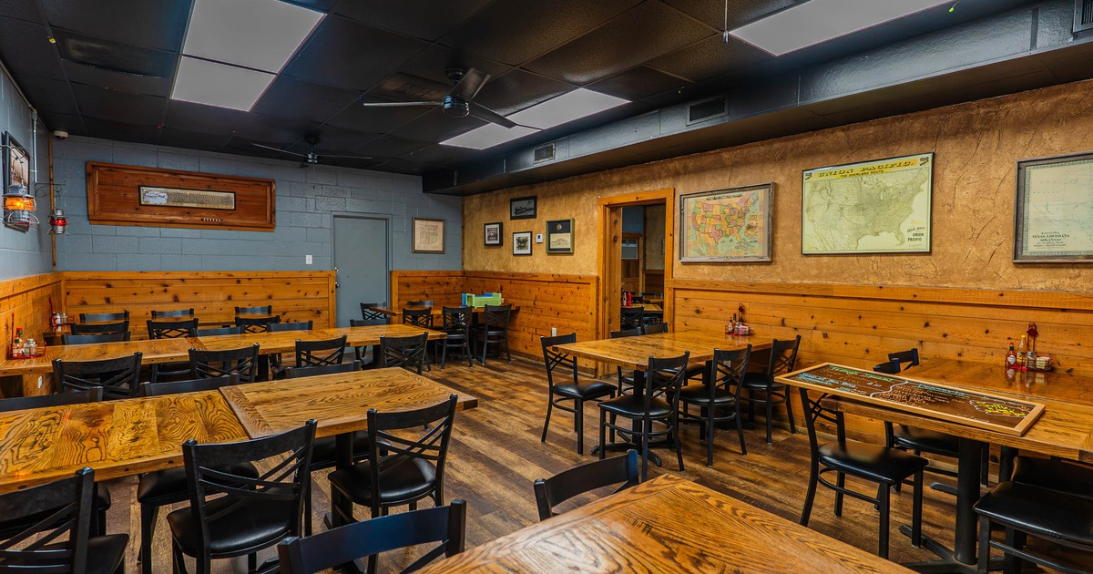 Interior of a rustic restaurant with wooden tables, black chairs, and framed maps on the walls