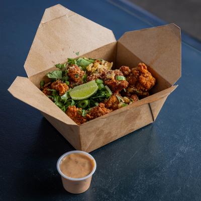 A paper box of crispy fried chicken nuggets sits on a dark table with a dipping sauce.
