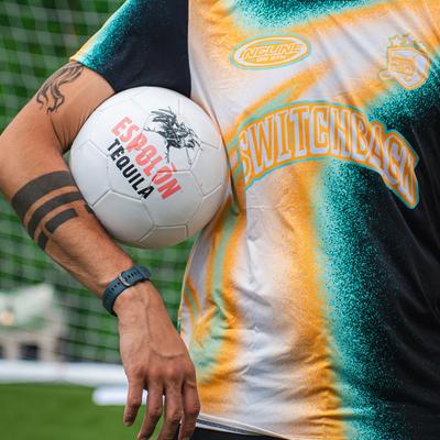 Person holding a branded soccer ball under their tattooed arm.