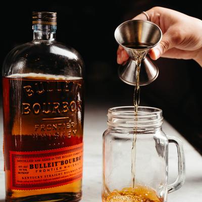A hand pouring a drink in a glass next to a bottle of bourbon.