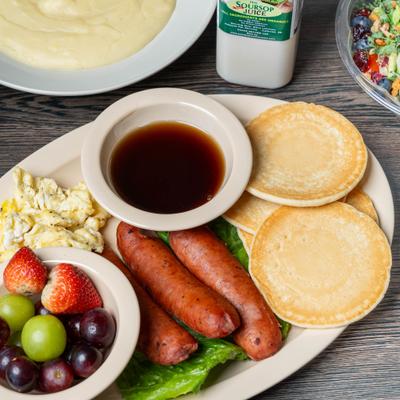 Pancakes with sausage, scrambled eggs, maple syrup, and fruit.