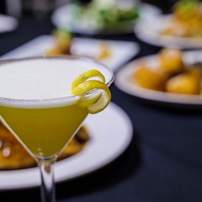 A close up of a yellow cocktail drink served in a Martini glass, various dishes in the background.