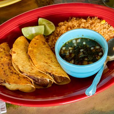 Birria tacos served with dipping broth, rice, and lime wedges on a red plate.