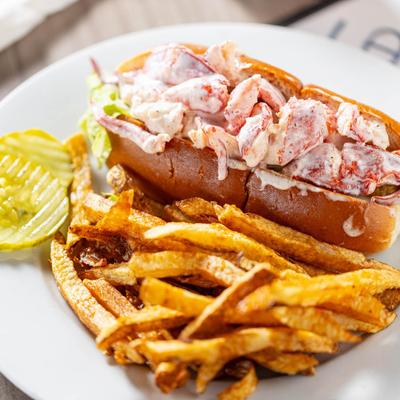 Lobster roll on a white plate with fries and pickle.