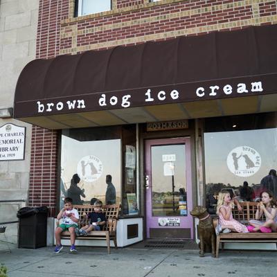Outdoor scene - two adults, two boys & two girls enjoying ice cream; a dog statue beside them.