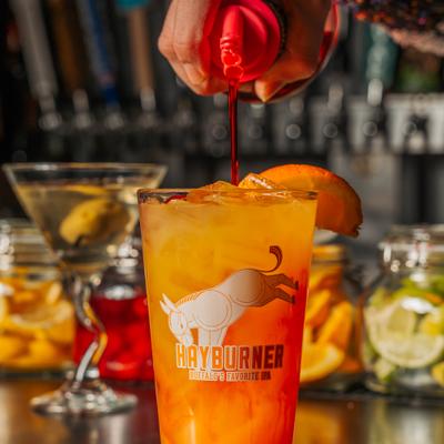 Bartender pouring red syrup into an orange cocktail garnished with an orange slice.