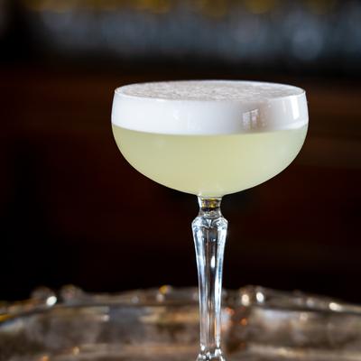Pale cocktail in a coupe glass with frothy top on a reflective bar.
