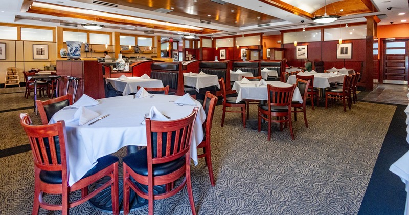 A restaurant interior with round tables, white tablecloths, wooden chairs, and warm lighting