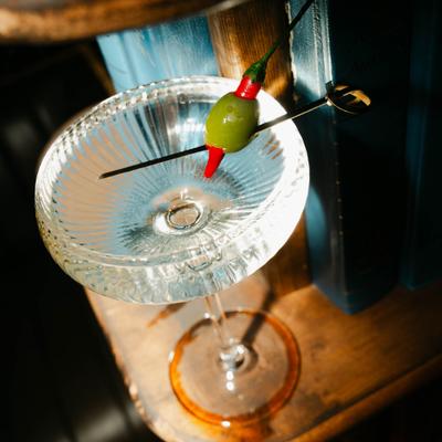 A clear cocktail glass sits on a wooden surface, with a green olive and red garnish on a skewer.