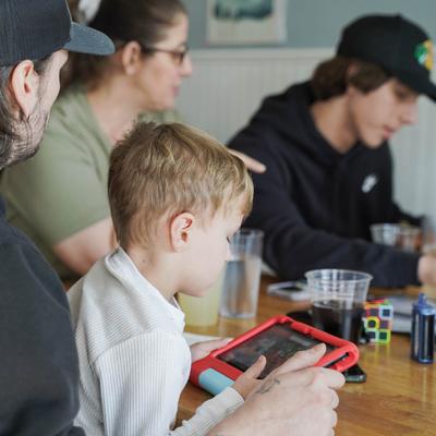 he child playing on a tablet at a table, surrounded by his family and drinks.