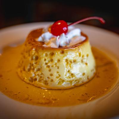 A flan rests on a white plate with caramel sauce, topped with whipped cream and a cherry.